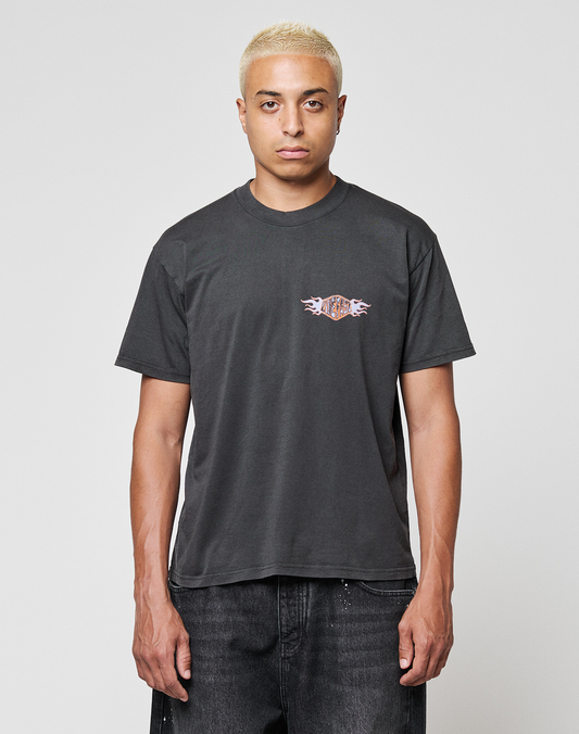 A person with short blond hair wears the LIVE FAST DIE YOUNG LFDY Fire Chest Tee, a black Regular Fit t-shirt featuring a small, colorful frontprint on the chest, paired with black jeans against a plain light gray background. | model