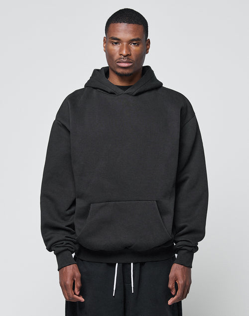 A man with a neutral expression stands against a plain light gray background, wearing LIVE FAST DIE YOUNG’s Basic Blank Hooded in an oversize fit black style and black pants with white drawstrings, his hands at his sides.