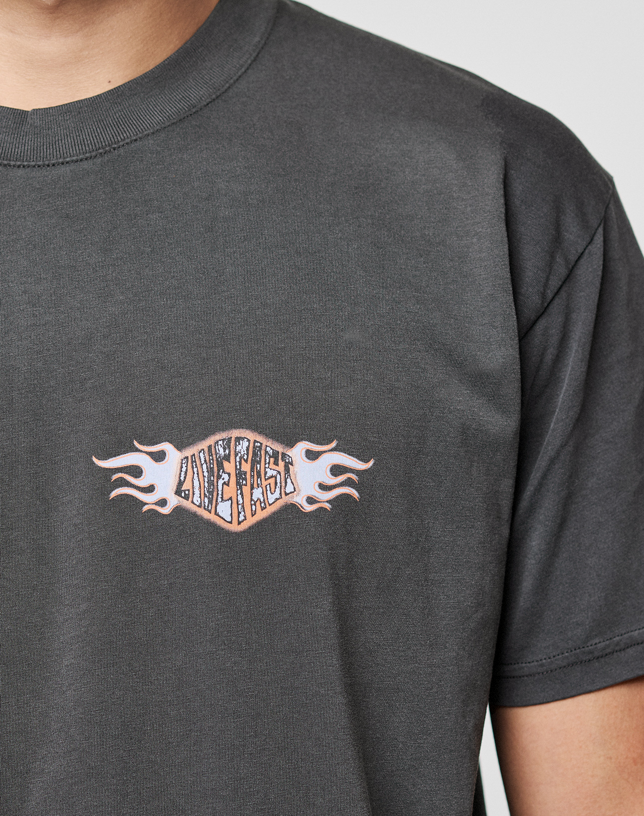 Close-up of a person in the LIVE FAST DIE YOUNG LFDY Fire Chest Tee, a dark gray 100% Baumwolle shirt with a small flame frontprint and “LIVE FAST” on the left chest. The face is partially out of frame.