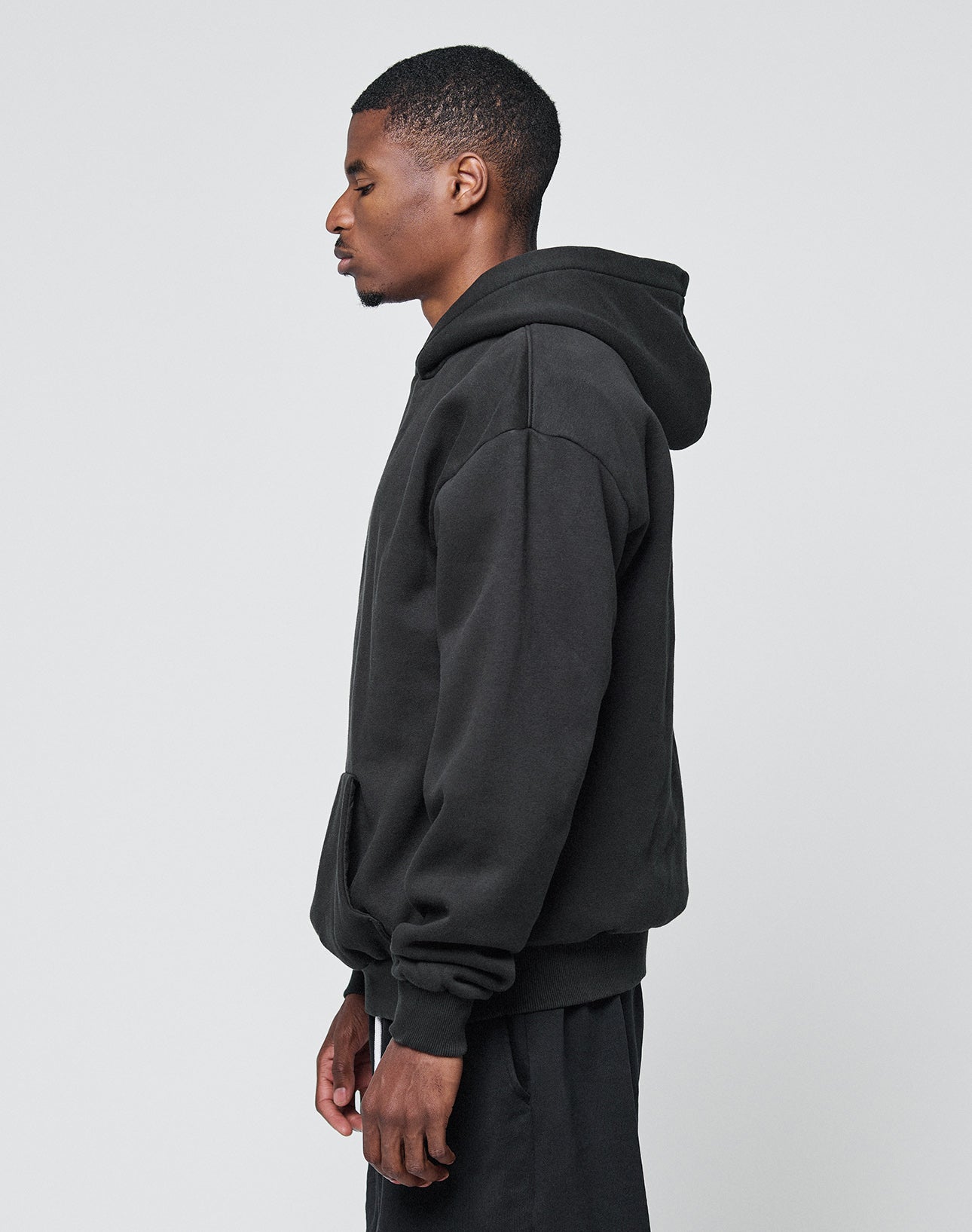 A man stands in profile against a light grey background, wearing the LIVE FAST DIE YOUNG Basic Blank Hooded—an oversize fit black hoodie made from a soft cotton-polyester blend—and black pants. His hands are relaxed by his sides, hood down.