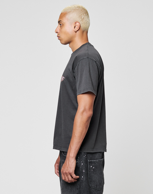 A person with short bleached blonde hair stands in profile, wearing the LIVE FAST DIE YOUNG LFDY Fire Chest Tee and black paint-splattered jeans, against a plain light gray background.
