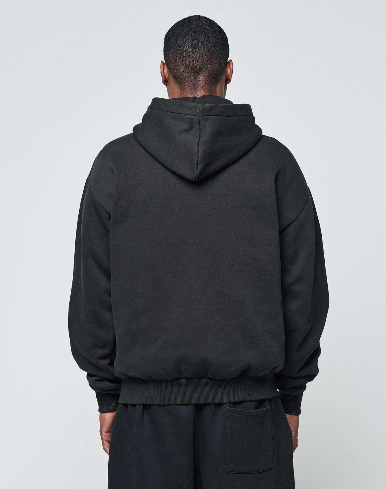 A person, back to camera, wears the LIVE FAST DIE YOUNG Basic Blank Hooded in an Oversize Fit with matching black pants made from a soft Baumwolle-Polyester blend, standing against a plain light gray background.