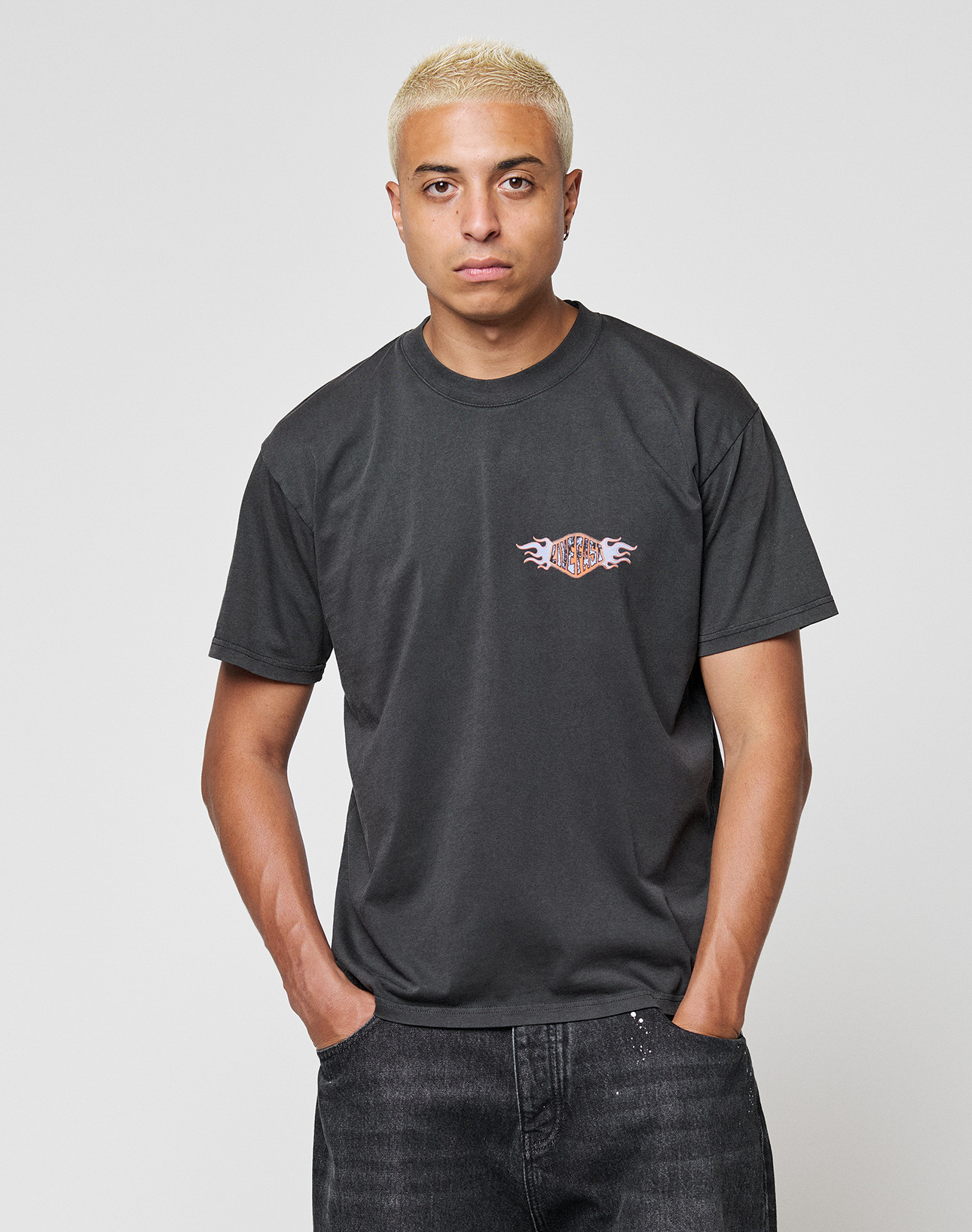 A person with short blond hair wears the LIVE FAST DIE YOUNG LFDY Fire Chest Tee, a regular fit black 100% cotton t-shirt with a small colorful chest frontprint, paired with dark jeans, standing against a plain light gray background.