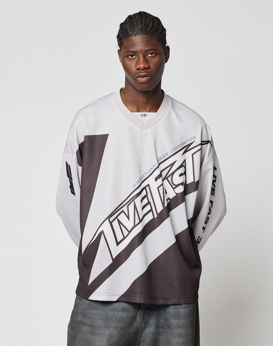 A young man with braids stands against a plain light background, wearing the oversize-fit LFDY Racetrack Jersey by LIVE FAST DIE YOUNG and casual dark pants. He gazes seriously at the camera with his hands behind his back.