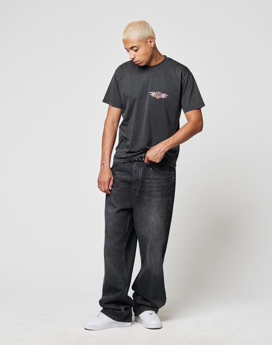 A person with short, bleached blonde hair wears the LIVE FAST DIE YOUNG LFDY Fire Chest Tee, loose-fitting dark jeans, and white sneakers, standing against a plain white background and looking down.