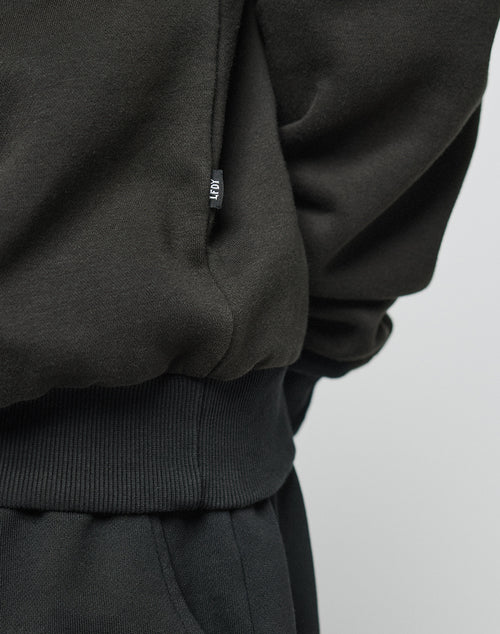 Close-up of a person wearing the LIVE FAST DIE YOUNG Basic Blank Hooded sweatshirt and matching pants in black, focusing on the waists small black tag with white text. The background is plain and light-colored.