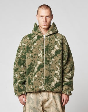 A person with short hair wears the LIVE FAST DIE YOUNG Live Fast Teddy Jacket—an oversize fit, fuzzy, camouflage-patterned piece with a hood and visor opening—paired with loose, light pants against a plain light background.