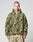 A person with short hair wears the LIVE FAST DIE YOUNG Live Fast Teddy Jacket—an oversize fit, fuzzy, camouflage-patterned piece with a hood and visor opening—paired with loose, light pants against a plain light background.
