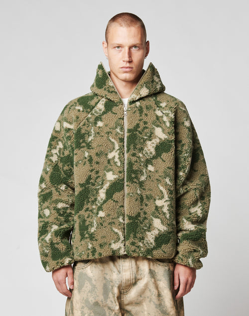 A person with short hair wears the LIVE FAST DIE YOUNG Live Fast Teddy Jacket—an oversize fit, fuzzy, camouflage-patterned piece with a hood and visor opening—paired with loose, light pants against a plain light background.