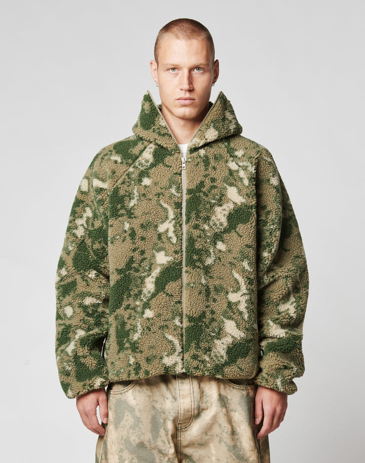A person with short hair wears the LIVE FAST DIE YOUNG Live Fast Teddy Jacket—an oversize fit, fuzzy, camouflage-patterned piece with a hood and visor opening—paired with loose, light pants against a plain light background. | model