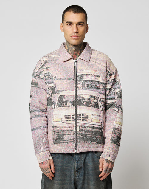 A man with short hair and tattoos stands facing forward, wearing the LIVE FAST DIE YOUNG LFDY Chase Jacket with a cars-in-traffic graphic print. He pairs it with loose jeans and poses against a plain white background.