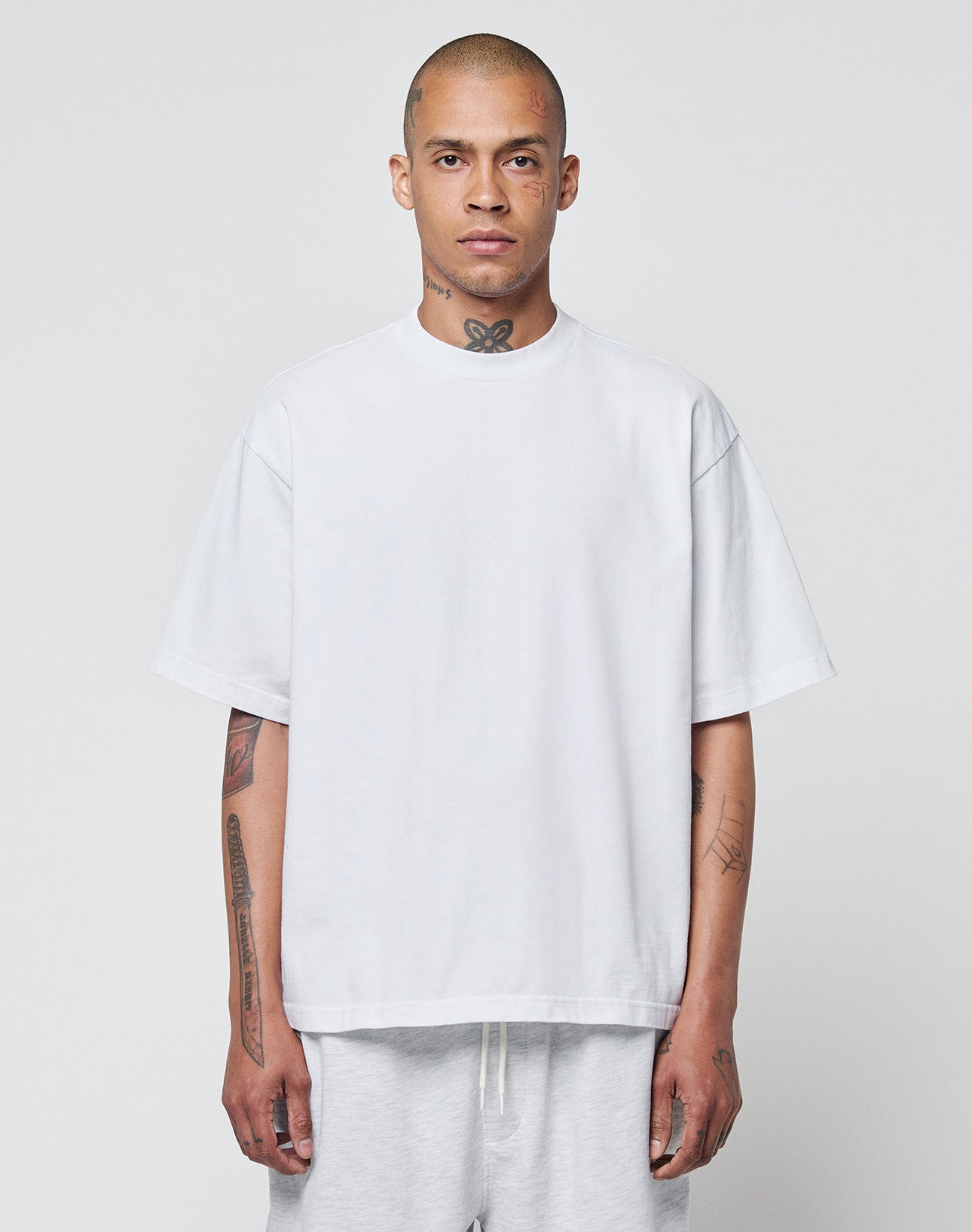 A man with a shaved head and tattoos wears the LIVE FAST DIE YOUNG Basic Heavy Tee, an oversize fit white t-shirt made from 100% cotton, paired with light pants. He stands against a plain light gray background, looking at the camera.
