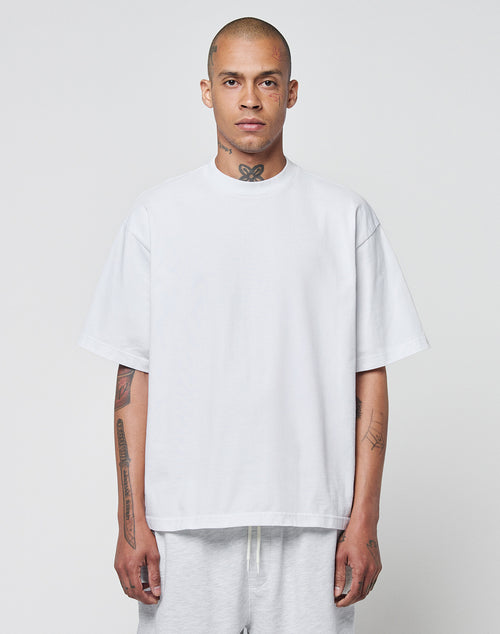 A man with a shaved head and tattoos wears the LIVE FAST DIE YOUNG Basic Heavy Tee, an oversize fit white t-shirt made from 100% cotton, paired with light pants. He stands against a plain light gray background, looking at the camera.