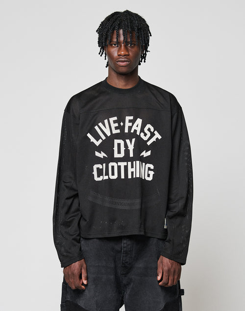 A young man with short dreadlocks models the LFDY Flash Jersey by LIVE FAST DIE YOUNG, an oversize black long-sleeve shirt with a bold white front print, standing against a plain light gray background.