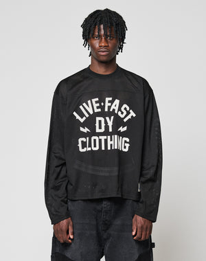 A young man with short dreadlocks models the LFDY Flash Jersey by LIVE FAST DIE YOUNG, an oversize black long-sleeve shirt with a bold white front print, standing against a plain light gray background.