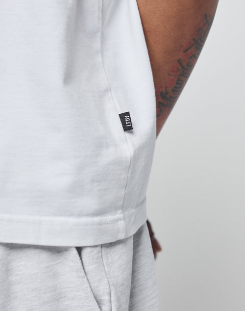 Close-up of a person in the LIVE FAST DIE YOUNG Basic Heavy Tee, an oversize fit white T-shirt made from 100% cotton, paired with light gray pants. A tattoo is on the arm, and a black fabric tag with white text is at the hem.