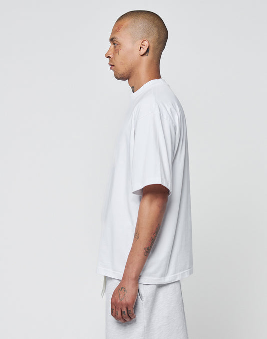 A tattooed, shaved-head person stands in profile against a plain background, wearing the LIVE FAST DIE YOUNG Basic Heavy Tee in oversized white 100% Baumwolle and matching white shorts.