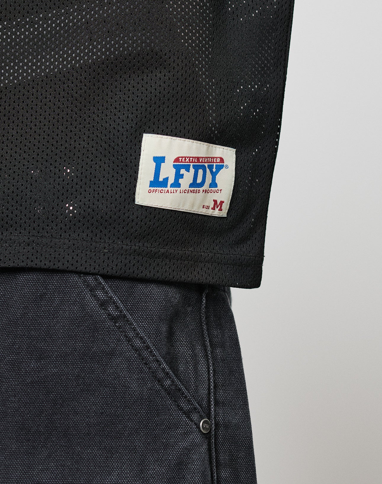 A close-up of the black mesh polyester LFDY Flash Jersey by LIVE FAST DIE YOUNG with a white label reading “LFDY Officially Licensed Product” in blue and red, shown next to dark gray denim pants.