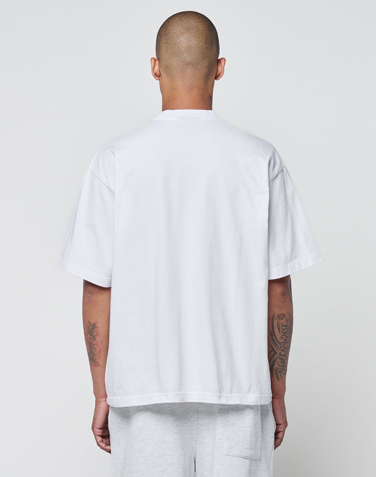 A person with a shaved head faces away, wearing a plain white Basic Heavy Tee by LIVE FAST DIE YOUNG, made of 100% Baumwolle, and light shorts. Tattoos show on both forearms against a plain light gray background.
