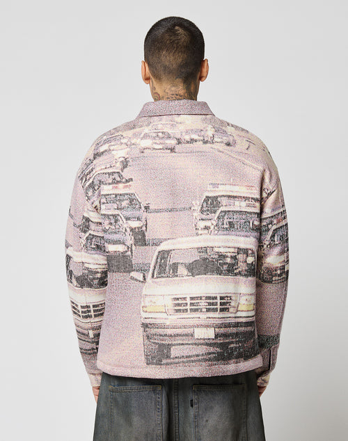 A person with short hair, seen from behind, wears the LIVE FAST DIE YOUNG LFDY Chase Jacket—an oversized Übergangsjacke with a graphic print of multiple cars in traffic. The plain background highlights the jacket’s striking design.