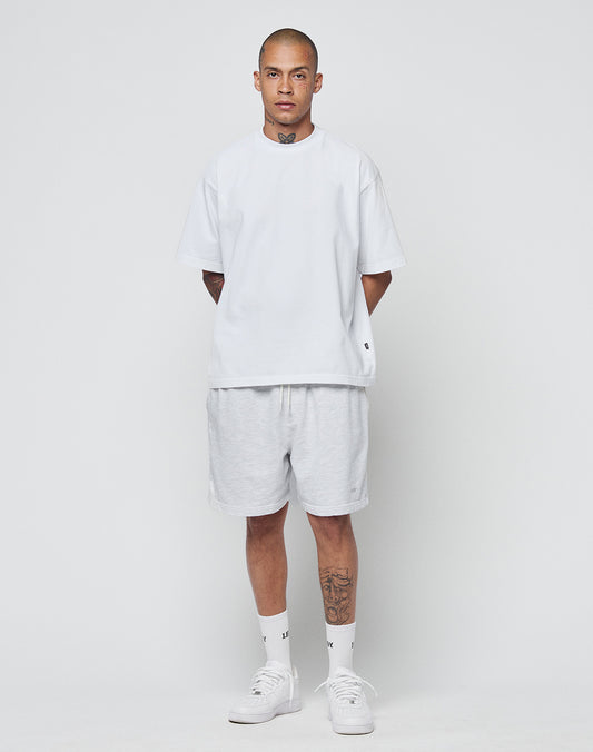 A person with short hair and visible tattoos models the LIVE FAST DIE YOUNG Basic Heavy Tee (100% Baumwolle) in an Oversize Fit, paired with light gray shorts, white socks, and sneakers against a plain white background.