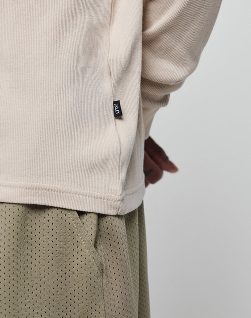 A close-up of a person wearing the LIVE FAST DIE YOUNG Basic Regular Rib Longsleeve in beige, featuring a small black fabric tag, paired with light olive green mesh pants. Only the torso and part of one hand are visible against a plain background.