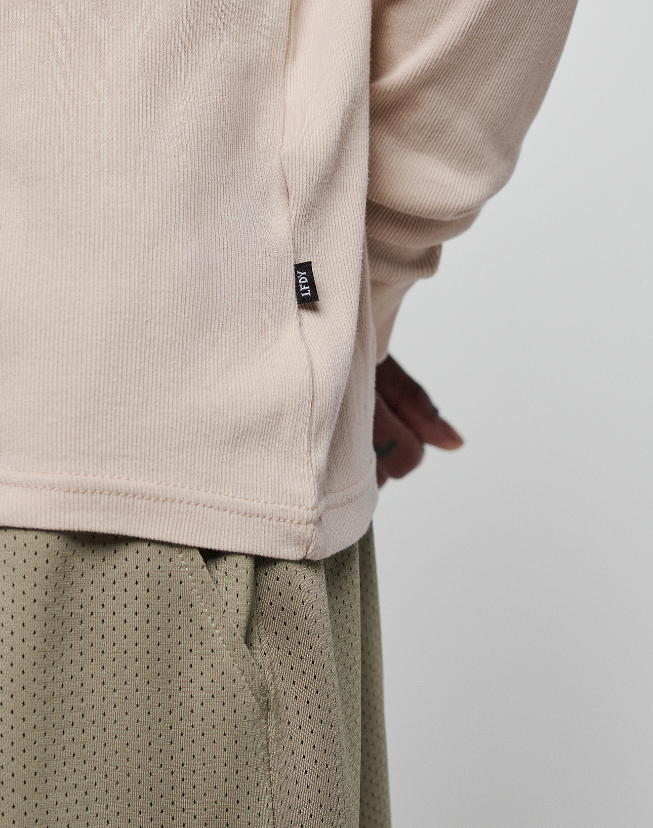 A close-up of a person wearing the LIVE FAST DIE YOUNG Basic Regular Rib Longsleeve in beige, featuring Ripp-Bündchen and a small black tag, paired with light olive green Regular Fit perforated pants. The person's hands are partially behind their back.
