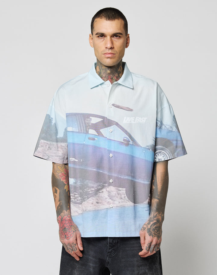 A tattooed person wears the LIVE FAST DIE YOUNG LFDY Crash Kid Shirt—an oversize, light blue short-sleeve button-up with a large blue car graphic and LIVE FAST text—standing against a plain white background.