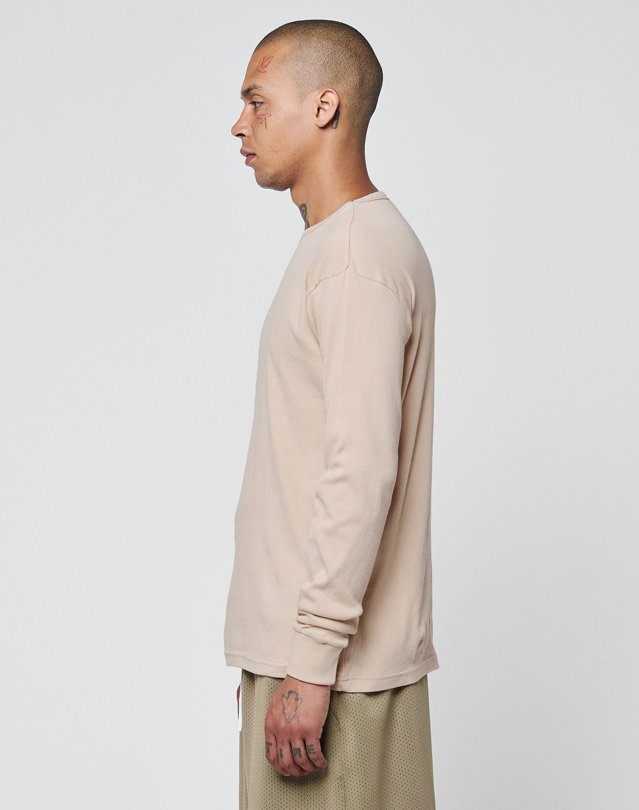 A person with a shaved head and tattoos stands in profile, wearing the beige LIVE FAST DIE YOUNG Basic Regular Rib Longsleeve and light-colored soft cotton pants, set against a plain white background.