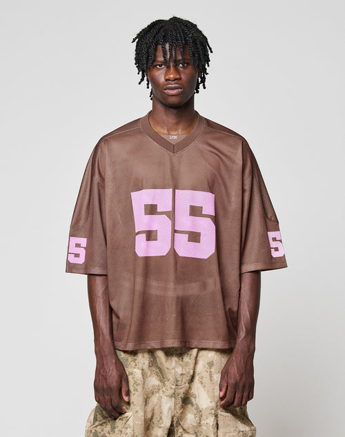 Someone with short, curly hair wears an oversized brown LFDY Mesh Jersey by LIVE FAST DIE YOUNG featuring a V-neck and pink 55 on the chest and sleeve, paired with beige camo pants, standing against a plain light background.