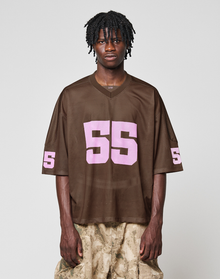 A person with short, twisted hair wears a loose brown LFDY Mesh Jersey by LIVE FAST DIE YOUNG with a pink 55 and V-neck, paired with light brown camo pants, standing against a plain light background.