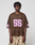A person with short, twisted hair wears a loose brown LFDY Mesh Jersey by LIVE FAST DIE YOUNG with a pink 55 and V-neck, paired with light brown camo pants, standing against a plain light background.