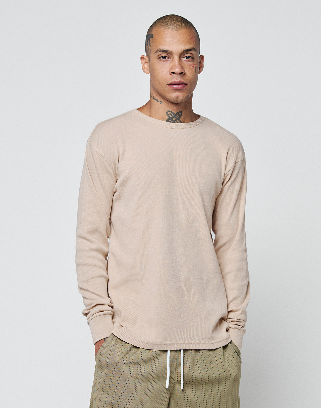 A person with a shaved head and neck tattoos stands against a white background, wearing the LIVE FAST DIE YOUNG Basic Regular Rib Longsleeve in beige with light brown drawstring pants for a comfortable regular fit look.