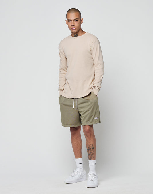 A person with short hair and a left-leg tattoo stands against a white background, wearing the LIVE FAST DIE YOUNG Basic Regular Rib Longsleeve in beige, olive green regular fit shorts, white crew socks, and white sneakers.