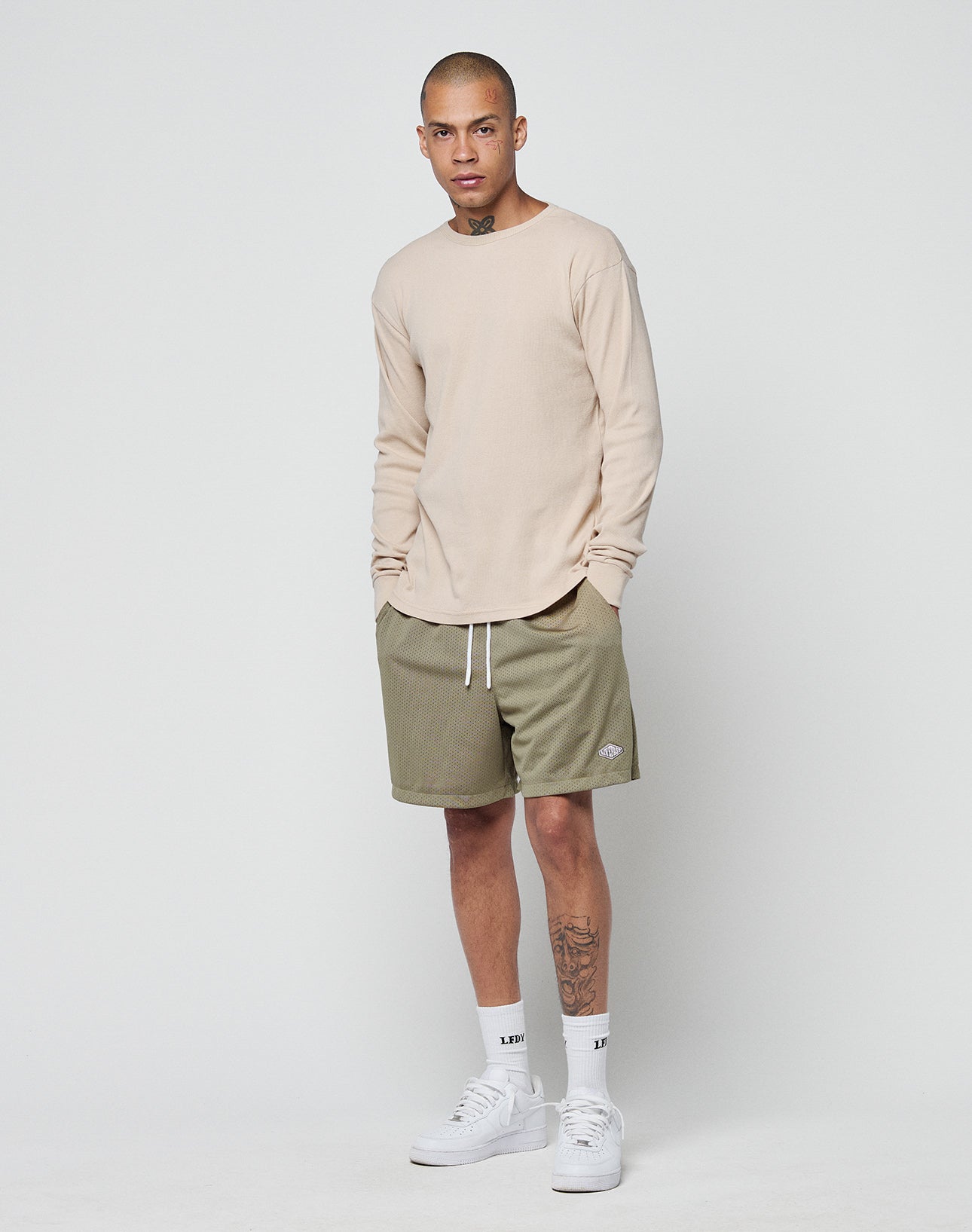 A person with short hair and a left-leg tattoo stands against a white background, wearing the LIVE FAST DIE YOUNG Basic Regular Rib Longsleeve in beige, olive green regular fit shorts, white crew socks, and white sneakers.