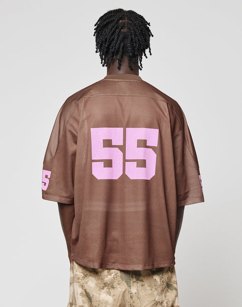 A person with short twists is seen from behind wearing the LIVE FAST DIE YOUNG LFDY Mesh Jersey in brown with a V-neck, oversize fit, and large pink number 55 on the back and sleeves, paired with beige patterned pants.