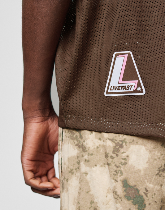 A close-up of a person wearing the LIVE FAST DIE YOUNG LFDY Mesh Jersey, an oversize brown mesh shirt with a pink and white L LIVEFAST patch, paired with camouflage pants. Their left arm hangs relaxed by their side.
