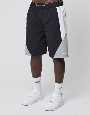 A person in black, gray, and white LIVE FAST DIE YOUNG Basic Nylon Trackshorts with elastic waistband and logo embroidery, white LFDY socks, and white sneakers stands against a plain white background.