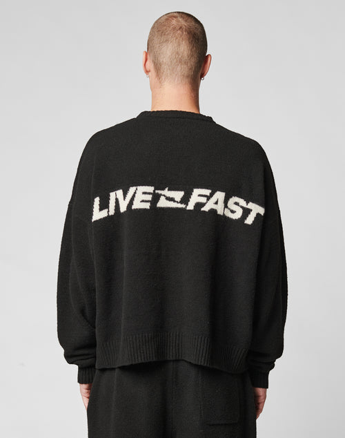 A person with short hair is seen from behind wearing the LIVE FAST DIE YOUNG Live Fast Boucle Sweater in black, featuring a white LIVE FAST slogan on the back, an oversize fit, and set against a plain light background.