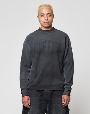 A person with short bleached blond hair wears the LIVE FAST DIE YOUNG LFDY Emblem Ghost Waffle Longsleeve, an oversized dark sweater with subtle lettering, and dark pants, standing facing forward against a plain light gray background.
