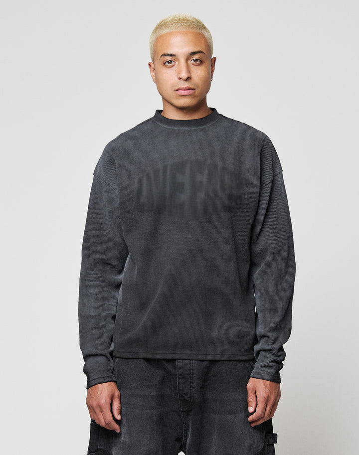 A person with short bleached blond hair wears the LIVE FAST DIE YOUNG LFDY Emblem Ghost Waffle Longsleeve, an oversized dark sweater with subtle lettering, and dark pants, standing facing forward against a plain light gray background. | model