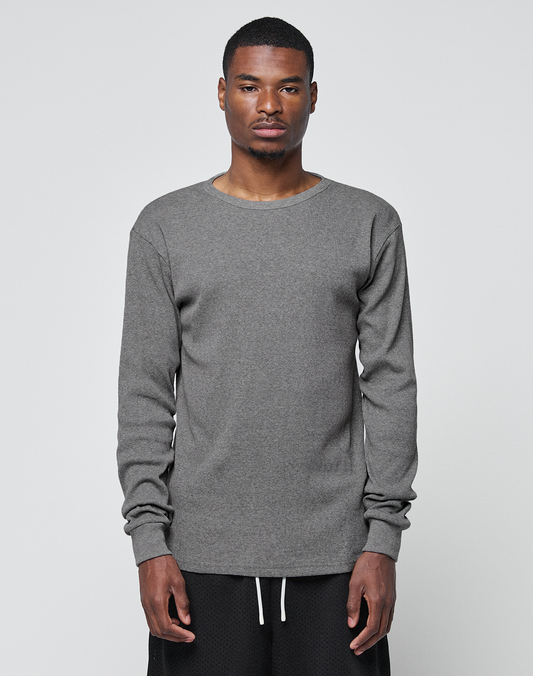 A man stands facing forward against a light gray background, wearing the LIVE FAST DIE YOUNG Basic Regular Rib Longsleeve and black pants, his neutral expression and relaxed arms emphasizing the regular fit style. | model