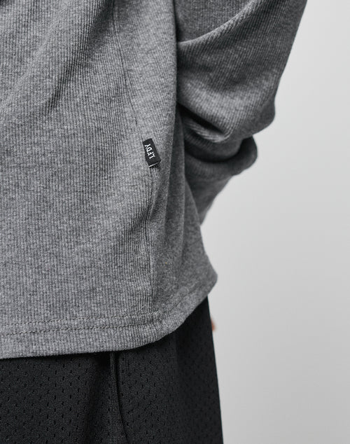 A close-up of a person wearing the LIVE FAST DIE YOUNG Basic Regular Rib Longsleeve in gray, featuring soft Baumwolle fabric and a small black ADT tag, paired with black mesh shorts against a plain light background.