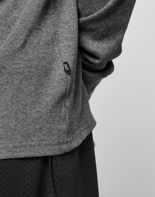 Close-up of a person wearing the LIVE FAST DIE YOUNG Basic Regular Rib Longsleeve in gray, featuring ribbed fabric, Ripp-Bündchen, and a small black ADT tag near the hem, paired with black textured pants. The arm is bent behind their back.