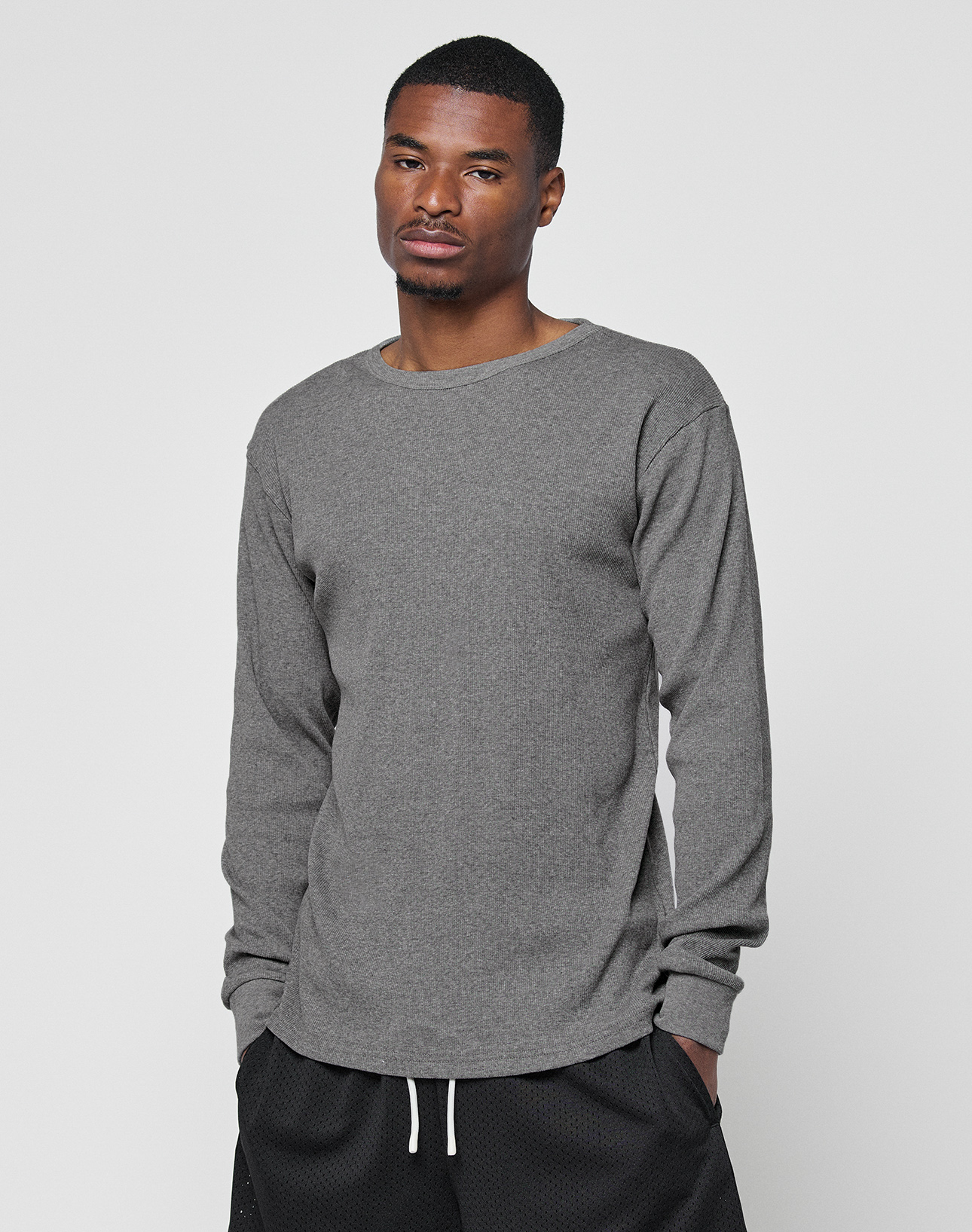 A man wears the LIVE FAST DIE YOUNG Basic Regular Rib Longsleeve in grey with Ripp-Bündchen and black shorts with white drawstrings, standing against a plain light background with his hands in his pockets.