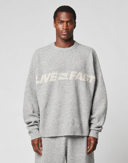 A person wears the LIVE FAST DIE YOUNG Live Fast Boucle Sweater in an oversized light gray fit with matching pants. The 100% polyester sweater displays blurred white LIVE FAST text across the chest. The background is plain and light-colored.