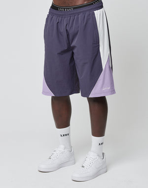 A person wearing LIVE FAST DIE YOUNG Basic Nylon Trackshorts in purple, white sports socks with LFDY text, and white sneakers stands against a plain white background. Only the lower body is visible.