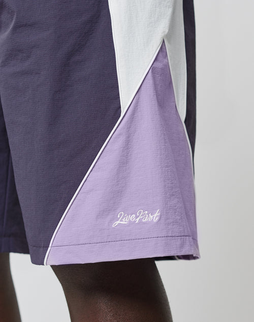 A close-up of a person wearing LIVE FAST DIE YOUNG Basic Nylon Trackshorts in a regular fit, color-blocked in purple, lavender, and white. Live Fast is embroidered on the lavender panel. Comfort is ensured by an elastic waistband; background is plain light.