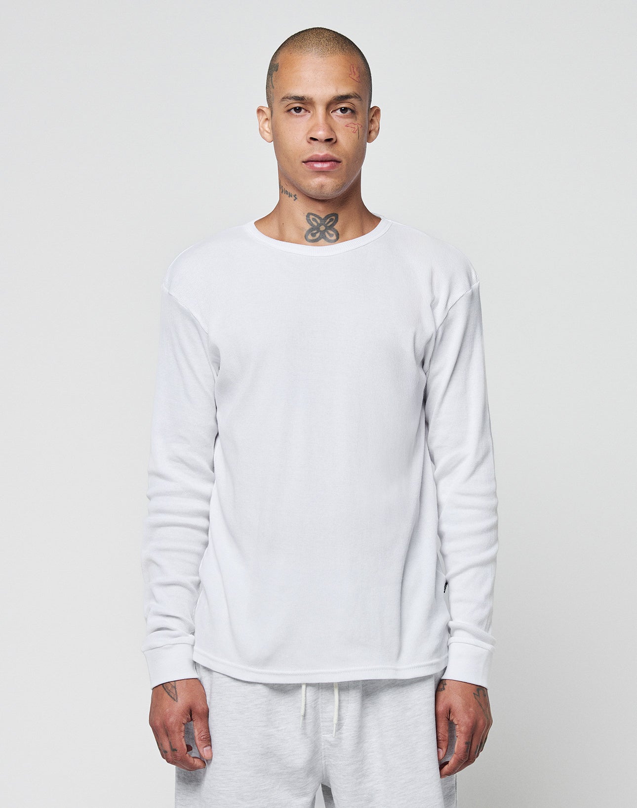 A person with a shaved head and neck tattoo wears the LIVE FAST DIE YOUNG Basic Regular Rib Longsleeve, a plain white shirt, along with matching white pants, standing against a light gray background and facing forward neutrally.