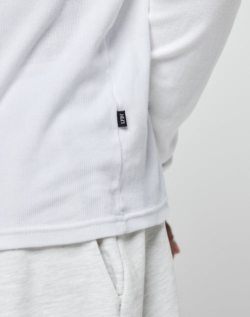 A close-up of a person wearing the LIVE FAST DIE YOUNG Basic Regular Rib Longsleeve in white and light grey sweatpants, featuring a small black EDIT tag on the shirts side seam. The persons hand rests by their side.